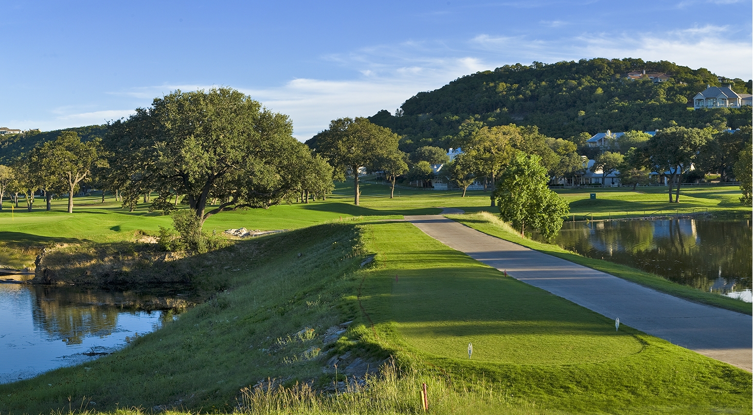 A golf course with a pond, green grass, and trees under a clear blue sky.