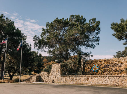 A stone wall entrance with trees and two flags waving in front.