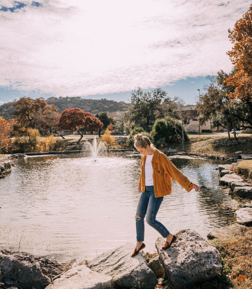 A person in a yellow jacket and jeans stepping on rocks by a pond. The trees in the background have autumn colors.