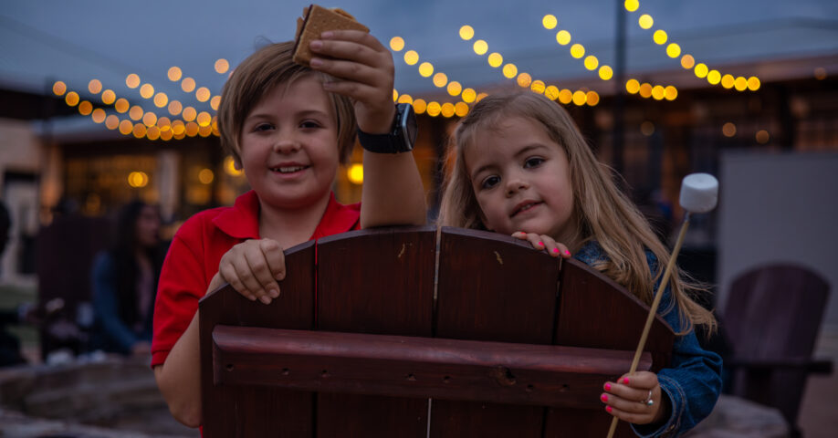two kids holding marshmallows near fire pit with string lights