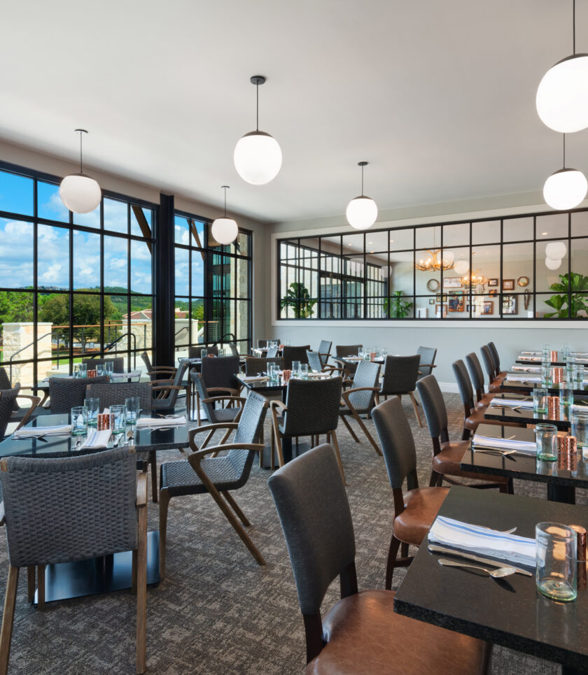 A brightly lit dining room with tables and chairs. There are large windows looking out to a green field and a blue sky.