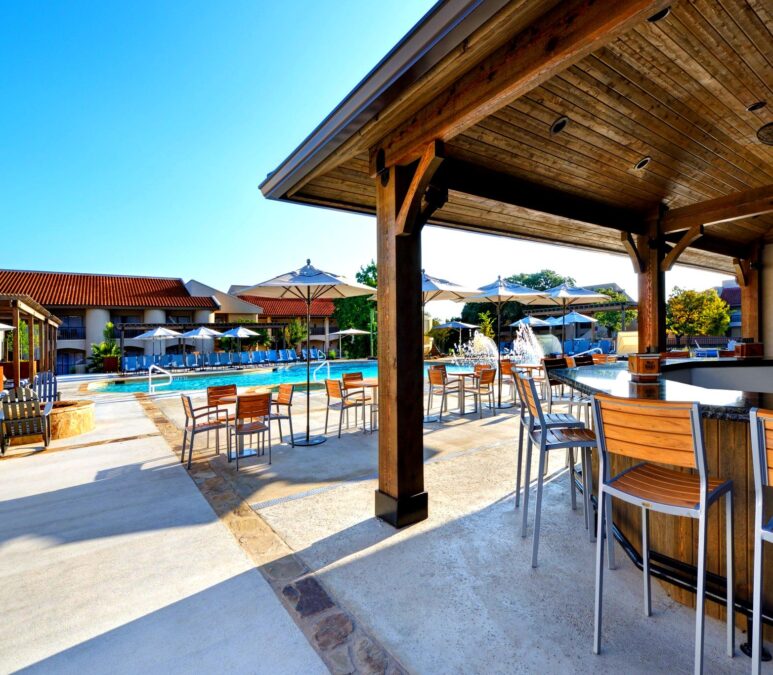 An outdoor bar with wooden stools and a covered patio, next to a pool area with lounge chairs.