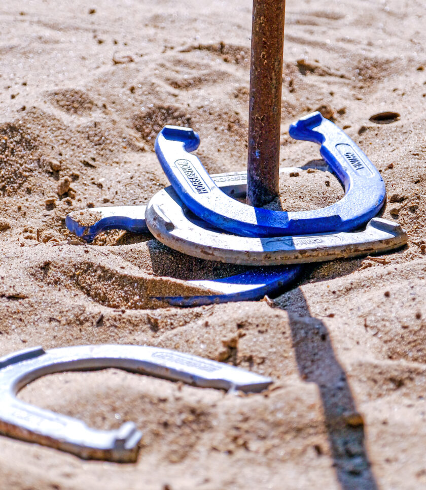 A blue horseshoe is looped around a metal stake in the sand, while a white horseshoe lies on the ground nearby.